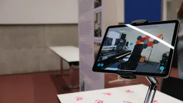 Ein Tablet steht auf einem Ständer und zeigt eine virtuelle Ansicht eines Raumes mit einem Klavier, Musikinstrumenten und einem Arbeitsbereich. Im Vordergrund sind mehrere rote, quadratische Markierungen auf einem Tisch zu sehen. Hintergrundbilder informieren über den Raum.