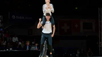 Ein männlicher Radfahrer fährt auf einem Fahrrad, während eine weibliche Tänzerin mit erhobenen Händen auf seinen Schultern steht. Beide sind in Sportkleidung und scheinen an einer sportlichen Darbietung teilzunehmen. Im Hintergrund sind Zuschauer und eine Wettkampfumgebung sichtbar.