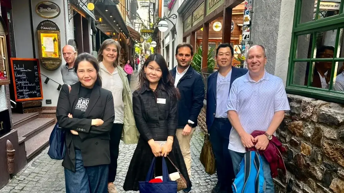 Eine Gruppe von sieben Personen steht in einer engen, gepflasterten Gasse. Sie lächeln und posieren für das Foto. Im Hintergrund sind mehrere Geschäfte und eine gemütliche Atmosphäre zu sehen. Die Menschen tragen legere Kleidung und scheinen sich wohl zu fühlen.