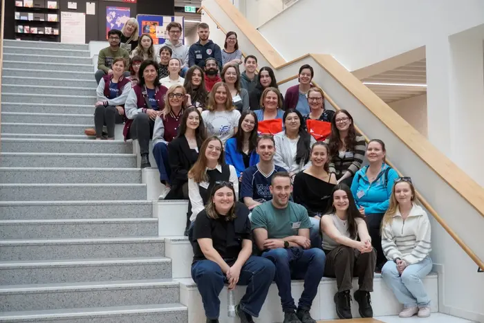 Eine Gruppe von etwa 40 Personen sitzt und steht auf einer Treppe in einem hellen Gebäude. Alle lächeln oder schauen freundlich in die Kamera. Die Anordnung der Personen zeigt eine diverse Gemeinschaft.