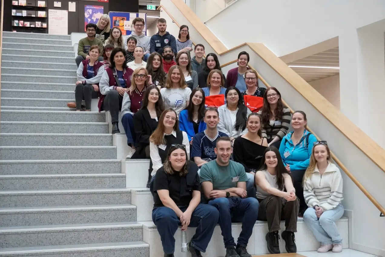 Eine Gruppe von etwa 40 Personen sitzt und steht auf einer Treppe in einem hellen Gebäude. Alle lächeln oder schauen freundlich in die Kamera. Die Anordnung der Personen zeigt eine diverse Gemeinschaft.