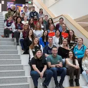 Eine Gruppe von etwa 40 Personen sitzt und steht auf einer Treppe in einem hellen Gebäude. Alle lächeln oder schauen freundlich in die Kamera. Die Anordnung der Personen zeigt eine diverse Gemeinschaft.