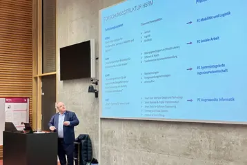 Vice President Prof. Dr. Andreas Brensing stands at a lectern and gives a presentation that can be seen on the screen behind him. He presents the research and transfer strategy of RheinMain University of Applied Sciences.