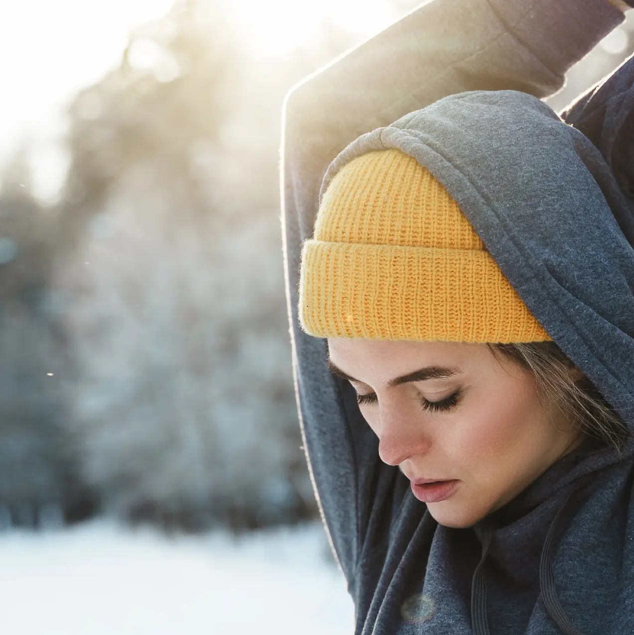 Eine junge Frau in einem grauen Hoodie und einer gelben Mütze dehnt sich im verschneiten Freien. Die Sonne scheint durch die Bäume im Hintergrund, während sie sich auf das Training vorbereitet.