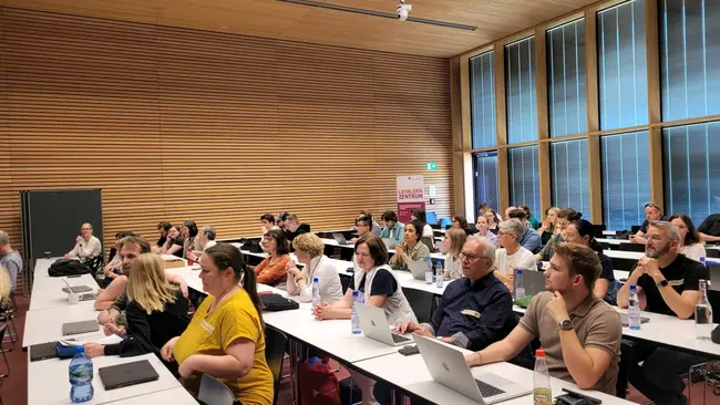 Eine Gruppe von Personen sitzt in einem Seminarraum an Tischen. Einige der Teilnehmenden schauen auf Laptops oder Notizblöcke. Der Raum hat große Fenster und eine Holzdecke, was eine helle und moderne Atmosphäre schafft.