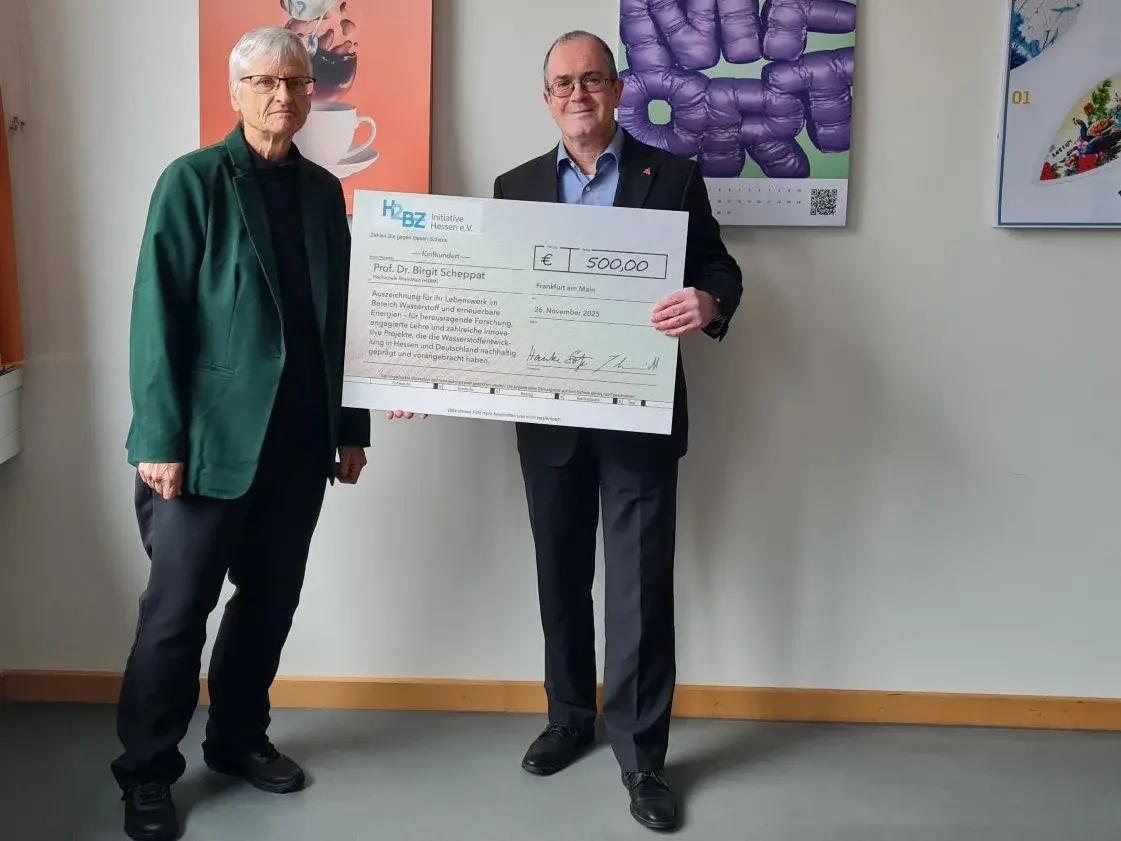 Zwei Personen stehen in einem Raum: Eine Frau mit grauen Haaren in einem schwarzen Oberteil und einer grünen Jacke, und ein Mann in einem blauen Sakko. Der Mann hält einen großen Scheck über 500 Euro, während die Frau neben ihm lächelt.