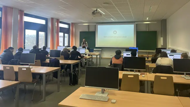 Ein Seminarraum mit mehreren Computern auf Tischen, in dem etwa zehn Schülerinnen und Schüler sitzen und auf einen Bildschirm schauen. Eine Lehrkraft steht vorne und präsentiert das KI-generierte Escapespiel zur Umweltbildung, während ein Projektor ein Bild an die Wand projiziert. Die Atmosphäre ist lernorientiert.