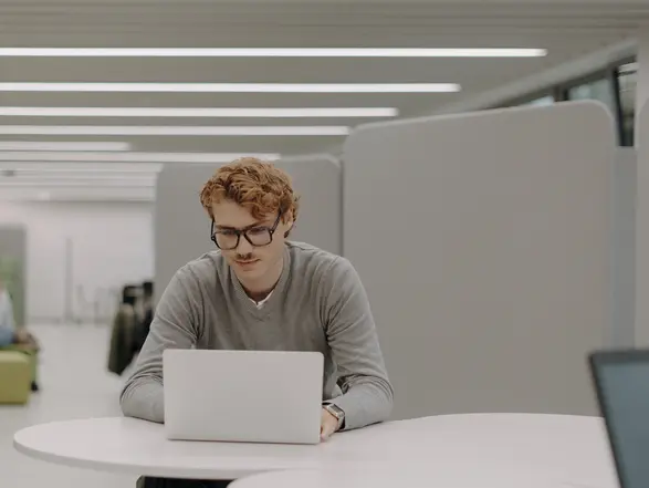 Ein junger Mann mit Brille sitzt an einem Tisch und arbeitet an einem Laptop. Er trägt einen grauen Pullover und konzentriert sich auf den Bildschirm. Im Hintergrund sind zwei Personen in einem entspannten Gespräch zu sehen, umgeben von modernen, grünen Möbeln in einem hellen Raum.