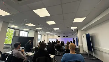 Eine Gruppe von Menschen sitzt in einem Konferenzraum und hört einem Vortrag zu. Im Hintergrund sind mehrere Personen auf der Bühne zu sehen, die eine Präsentation halten. Die Atmosphäre wirkt konzentriert und professionell.