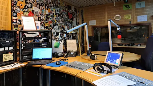 Ein Radio-Studium mit einem Mischpult, Mikrofonen und einem Computer. An der Wand hängen viele Fotos und Notizen. Auf dem Tisch liegen Kopfhörer und ein Dokument mit Text. Die Atmosphäre wirkt kreativ und einladend für die Produktion von Sendungen.