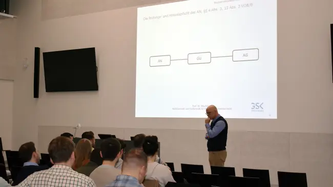 Prof. Oliver Moufang von GSK Stockmann steht vor einem Publikum und erläutert Inhalte, die auf einer Präsentationsfolie dargestellt sind beim Wiesbadener Baubetriebsabend an der Hochschule RheinMain.