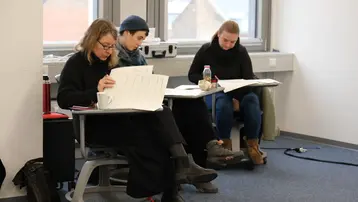 Drei Personen sitzen in einem Raum. Zwei Frauen konzentrieren sich auf Unterlagen, während eine dritte Frau entspannt zuschaut. Alle tragen dunkle Kleidung. Der Raum ist hell erleuchtet und hat große Fenster mit einem Blick nach draußen.