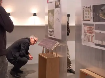 Eine Person beugt sich vor, um ein Architekturmodell in einer Ausstellung zu betrachten. Im Hintergrund sind Plakate mit Entwürfen und Informationen zu sehen.