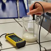 Eine Hand hält einen Messfühler, der in ein Becherglas mit einer weißen Flüssigkeit eingetaucht ist. Neben dem Becherglas befindet sich ein gelbes Messgerät. Das Bild zeigt eine Laborsituation zur chemischen Analyse oder Messung.