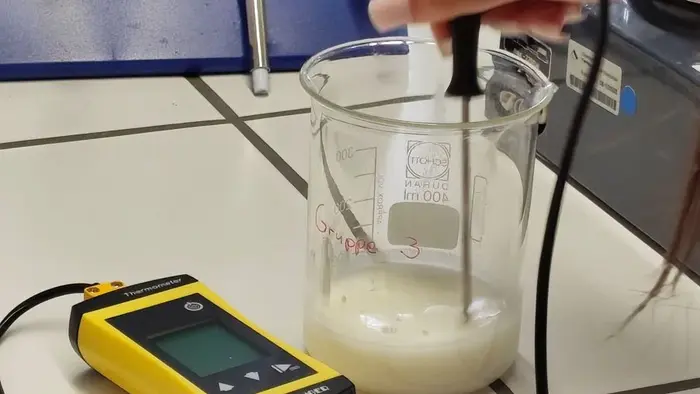 Eine Hand hält einen Messfühler, der in ein Becherglas mit einer weißen Flüssigkeit eingetaucht ist. Neben dem Becherglas befindet sich ein gelbes Messgerät. Das Bild zeigt eine Laborsituation zur chemischen Analyse oder Messung.
