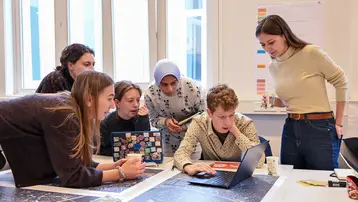 A group of six students are standing around a table with maps and a laptop on it. They are clearly discussing a topic while one of them works on the laptop. The atmosphere is enthusiastic and cooperative.