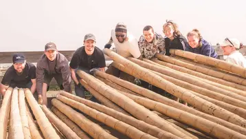 Gruppenszene von acht Personen, die zusammen große Holzbalken halten. Die Menschen lächeln und arbeiten gemeinsam. Im Hintergrund ist eine andere Struktur zu erkennen, die das Team bei ihrer Tätigkeit unterstützt. Es herrscht eine freundliche, kooperative Atmosphäre.