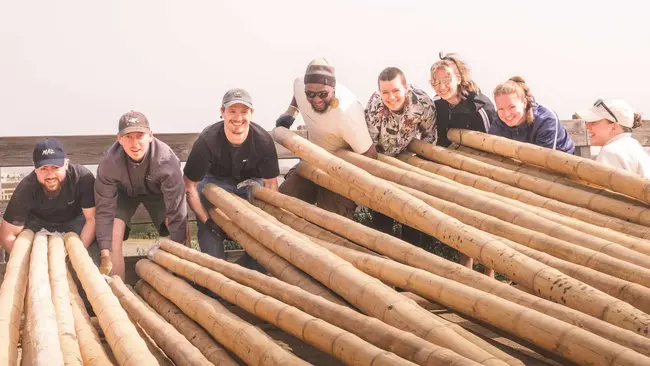 Gruppenszene von acht Personen, die zusammen große Holzbalken halten. Die Menschen lächeln und arbeiten gemeinsam. Im Hintergrund ist eine andere Struktur zu erkennen, die das Team bei ihrer Tätigkeit unterstützt. Es herrscht eine freundliche, kooperative Atmosphäre.