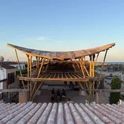 Eine Holzstruktur mit einem geschwungenen Dach steht über einem offenen Raum, umgeben von einem bepflanzten Bereich und Sand. Im Hintergrund ist das Meer zu sehen, während einige Personen im unteren Bereich sitzen. Der Himmel zeigt einen sanften Farbverlauf des Sonnenuntergangs.