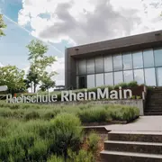 Das Bild zeigt das Gebäude der Hochschule RheinMain, umgeben von grünen Pflanzen und Sträuchern. Im Vordergrund steht der Schriftzug "Hochschule RheinMain". Der Himmel ist teilweise bewölkt, und das Gesamtbild vermittelt einen einladenden Eindruck.