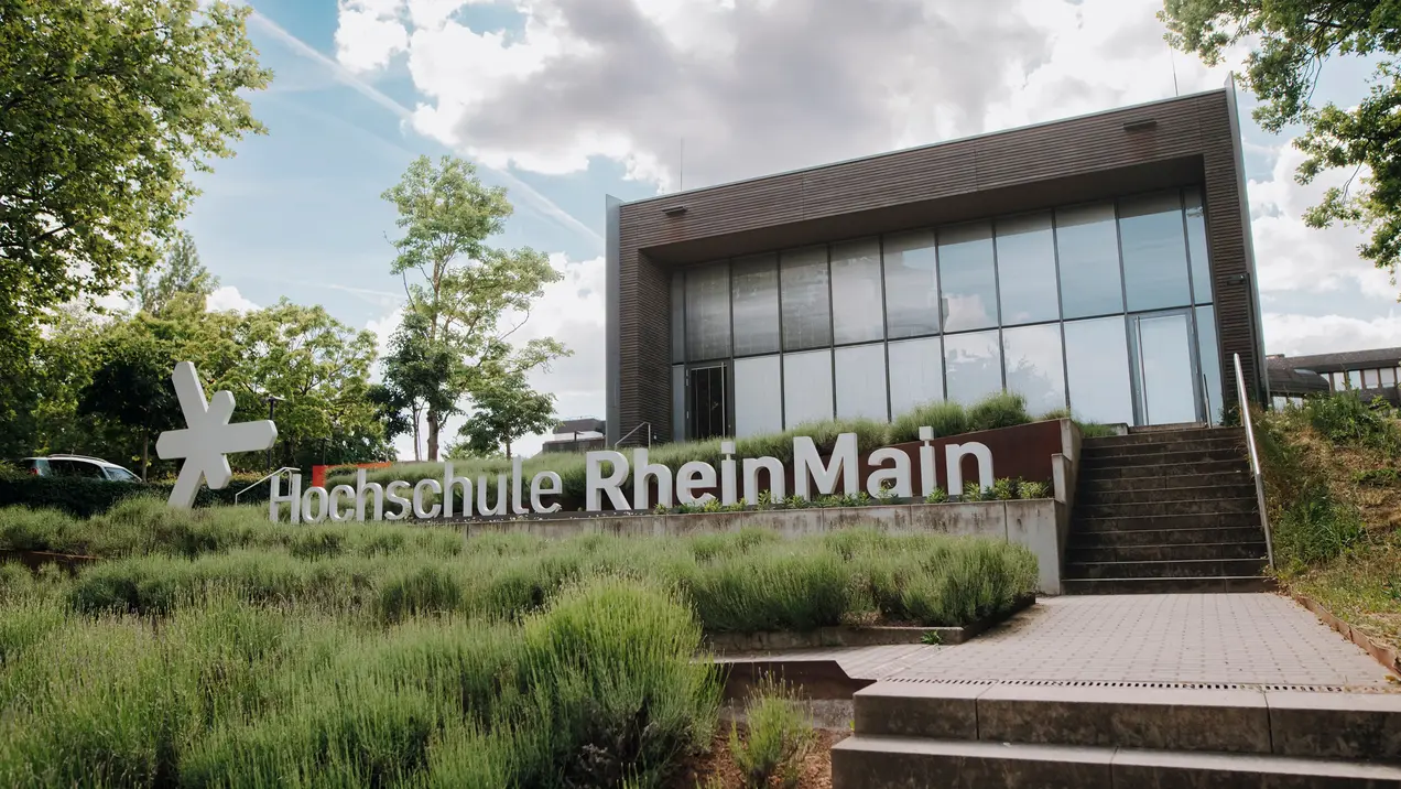 Das Bild zeigt das Gebäude der Hochschule RheinMain, umgeben von grünen Pflanzen und Sträuchern. Im Vordergrund steht der Schriftzug "Hochschule RheinMain". Der Himmel ist teilweise bewölkt, und das Gesamtbild vermittelt einen einladenden Eindruck.