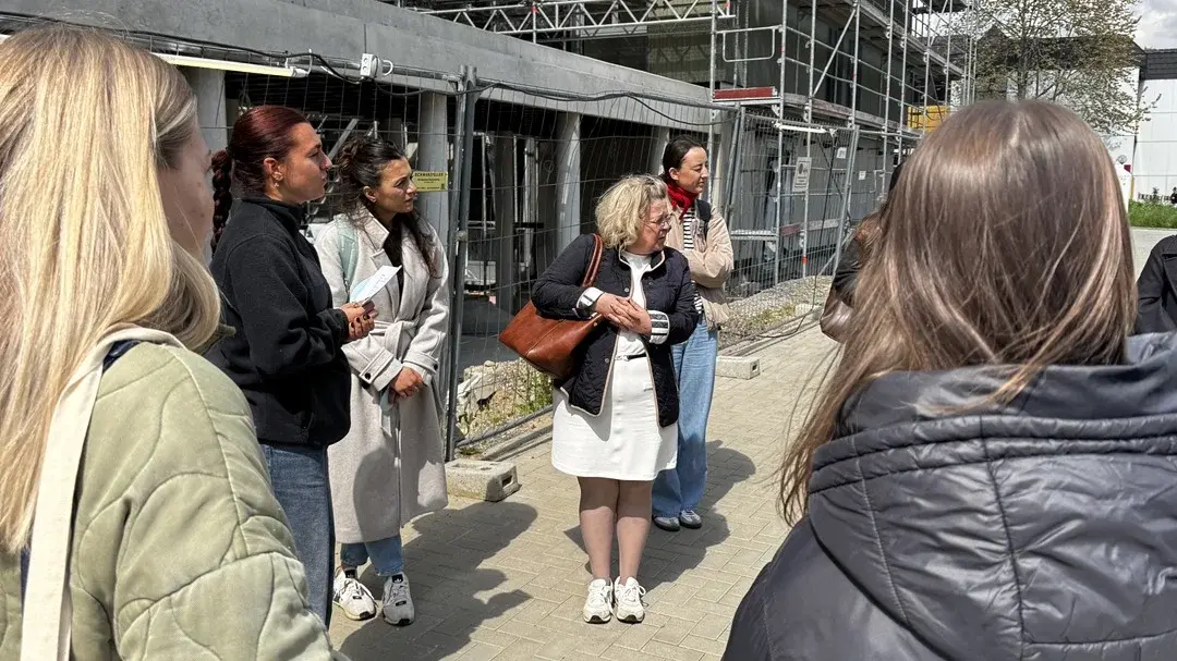 Eine Gruppe von sechs Personen steht im Freien vor einem unvollendeten Gebäude, das von Gerüsten umgeben ist. Eine Frau in der Mitte spricht, während die anderen aufmerksam zuhören. Es ist sonnig, und die Atmosphäre wirkt informativ und freundlich.