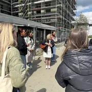 Eine Gruppe von sechs Personen steht im Freien vor einem unvollendeten Gebäude, das von Gerüsten umgeben ist. Eine Frau in der Mitte spricht, während die anderen aufmerksam zuhören. Es ist sonnig, und die Atmosphäre wirkt informativ und freundlich.