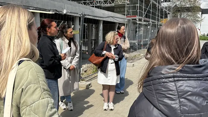 Eine Gruppe von sechs Personen steht im Freien vor einem unvollendeten Gebäude, das von Gerüsten umgeben ist. Eine Frau in der Mitte spricht, während die anderen aufmerksam zuhören. Es ist sonnig, und die Atmosphäre wirkt informativ und freundlich.