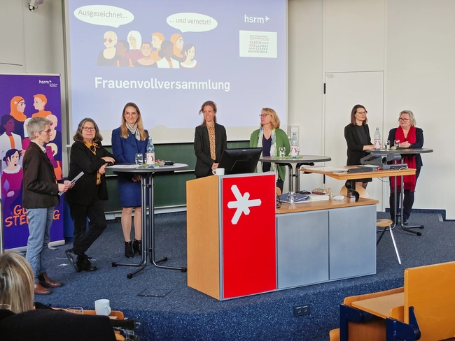 Die Veranstaltung zeigt eine Frauenvollversammlung mit sieben Rednerinnen auf der Bühne. Im Hintergrund sind Präsentationen zu sehen, die auf die Themen "Ausgezeichnet... und vernetzt!" hinweisen. Die Atmosphäre ist professionell und geprägt von engagierten Gesprächen.