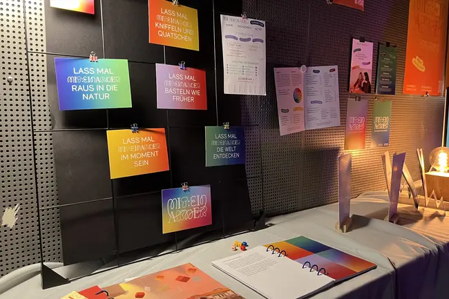 Ein Ausstellungsstand mit bunten Postkarten. Darunter liegt ein offenes Notizbuch und Farbproben.