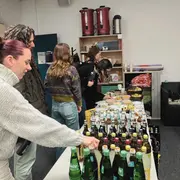 Eine Gruppe von vier Personen steht um einen Tisch, der mit verschiedenen Getränken und Snacks gedeckt ist. Im Vordergrund greift eine Person mit kurzen, bunten Haaren nach einer Flasche. Im Hintergrund sind weitere Personen, die sich ebenfalls um das Buffet kümmern.