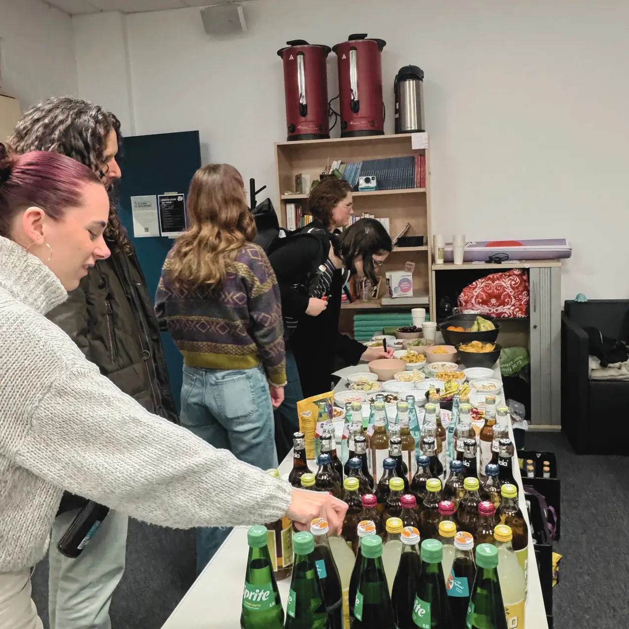 Eine Gruppe von vier Personen steht um einen Tisch, der mit verschiedenen Getränken und Snacks gedeckt ist. Im Vordergrund greift eine Person mit kurzen, bunten Haaren nach einer Flasche. Im Hintergrund sind weitere Personen, die sich ebenfalls um das Buffet kümmern.