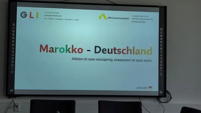 Auf einem Bildschirm steht der Text: "Marokko – Deutschland" in bunten Buchstaben. Darunter steht: "Alleine ist man einzigartig, zusammen ist man stark." Das Datum "23.03.2023" ist ebenfalls zu sehen. Der Kontext scheint eine Präsentation oder ein Meeting zu sein.
