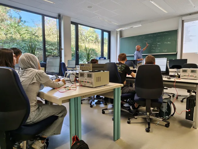 Ein Lehrer steht an einem Tisch und erklärt etwas an einer Tafel in einem Klassenzimmer. Acht Studierende sitzen an Tischen mit Computern und Messgeräten. Große Fenster zeigen einen Garten im Hintergrund. Das Bild vermittelt eine Lernumgebung in der Technik oder Naturwissenschaften.