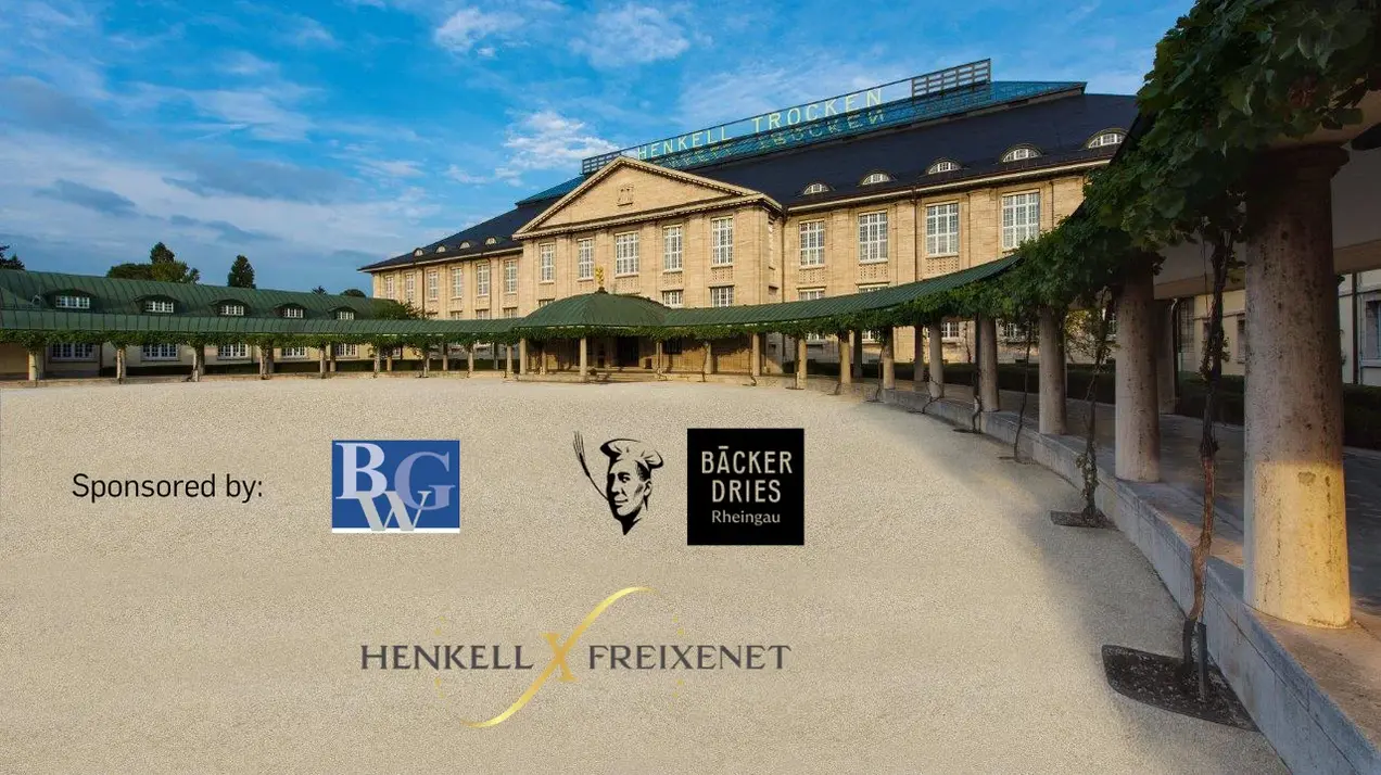Das Bild zeigt ein historisches Gebäude mit grünen Dächern und großen Fenstern, umgeben von einer Sandfläche. Oben auf dem Gebäude steht "HENKELL TROCKEN". Im Vordergrund sind die Logos von Henkell, Freixenet und Sponsoren zu sehen. Der Himmel ist blau mit einigen Wolken.