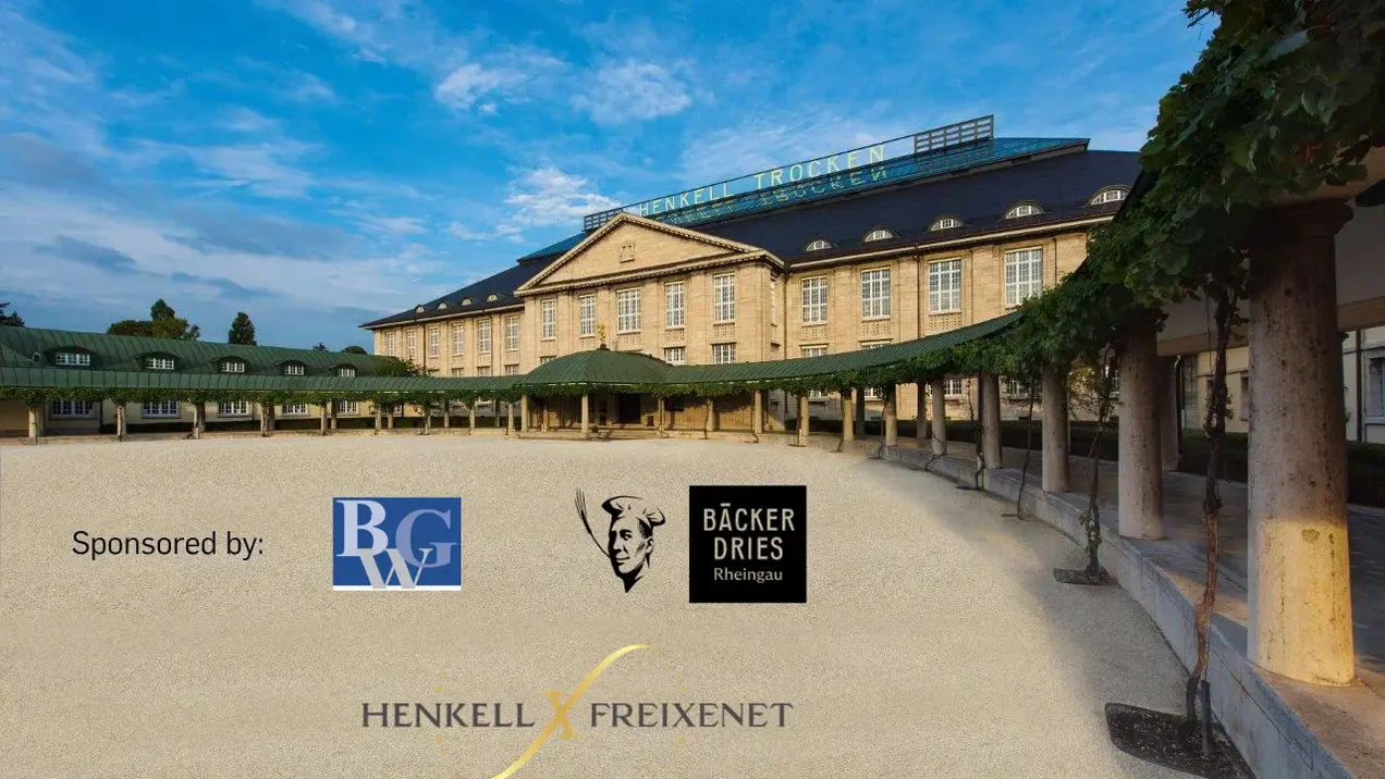 Das Bild zeigt ein historisches Gebäude mit grünen Dächern und großen Fenstern, umgeben von einer Sandfläche. Oben auf dem Gebäude steht "HENKELL TROCKEN". Im Vordergrund sind die Logos von Henkell, Freixenet und Sponsoren zu sehen. Der Himmel ist blau mit einigen Wolken.