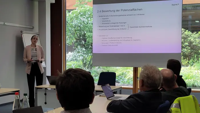 Eine Präsentatorin steht vor einem Bildschirm, auf dem der Text "2.4 Bewertung der Potenzialflächen" zu sehen ist. Im Hintergrund sitzen mehrere Personen, die aufmerksam zuhören. Der Raum ist hell und bietet einen Blick auf das Grün außerhalb.