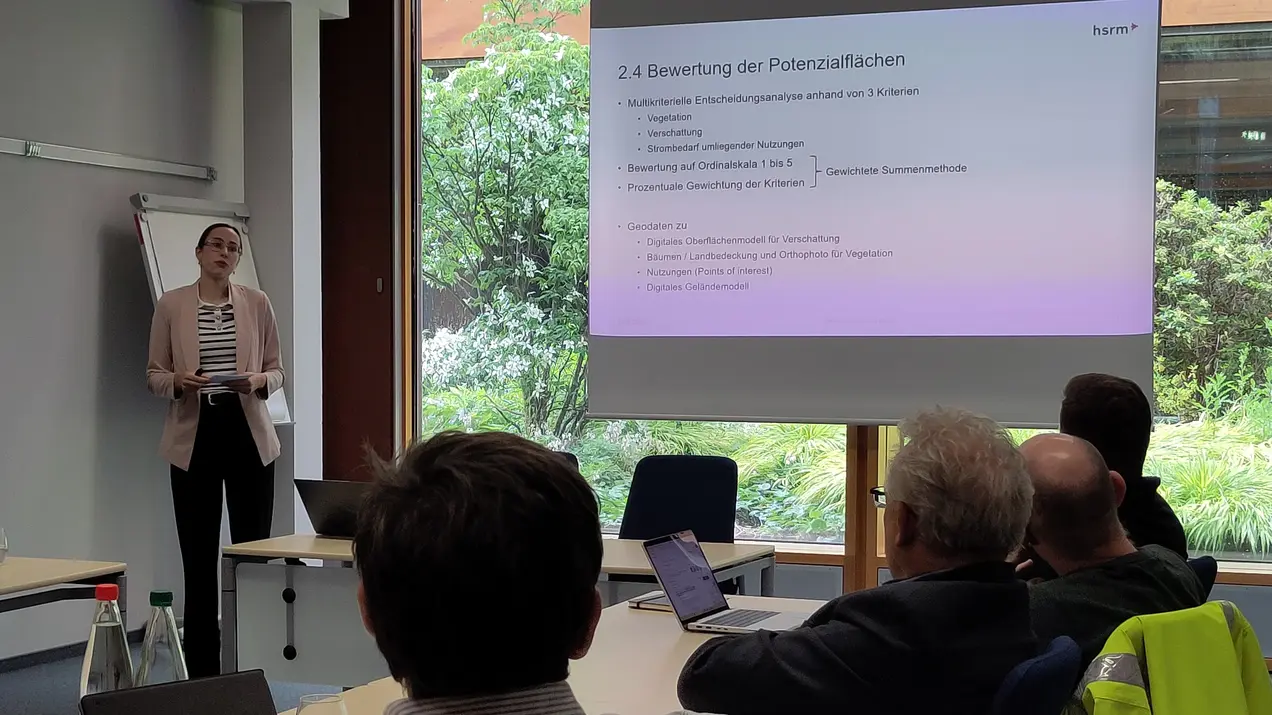 Eine Präsentatorin steht vor einem Bildschirm, auf dem der Text "2.4 Bewertung der Potenzialflächen" zu sehen ist. Im Hintergrund sitzen mehrere Personen, die aufmerksam zuhören. Der Raum ist hell und bietet einen Blick auf das Grün außerhalb.