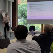 Eine Präsentatorin steht vor einem Bildschirm, auf dem der Text "2.4 Bewertung der Potenzialflächen" zu sehen ist. Im Hintergrund sitzen mehrere Personen, die aufmerksam zuhören. Der Raum ist hell und bietet einen Blick auf das Grün außerhalb.