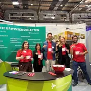 Eine Gruppe von fünf Personen steht an einem Messestand der Hochschule RheinMain. Alle tragen rote T-Shirts und halten Broschüren oder Kärtchen in der Hand. Der Stand ist mit Informationen über Ingenieurwissenschaften dekoriert, und im Hintergrund sind technische Geräte sichtbar.