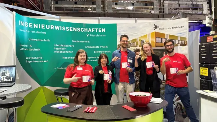 Eine Gruppe von fünf Personen steht an einem Messestand der Hochschule RheinMain. Alle tragen rote T-Shirts und halten Broschüren oder Kärtchen in der Hand. Der Stand ist mit Informationen über Ingenieurwissenschaften dekoriert, und im Hintergrund sind technische Geräte sichtbar.
