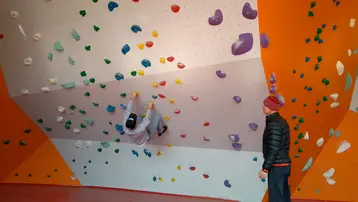 Ein Kletterer übt an einer bunten Bouldering-Wand. Er hängt kopfüber und nutzt verschiedene farbige Griffe. Eine Person in schwarzer Jacke schaut ihm aufmerksam zu. Die Wand ist orange und mit zahlreichen Griffen in verschiedenen Farben ausgestattet.