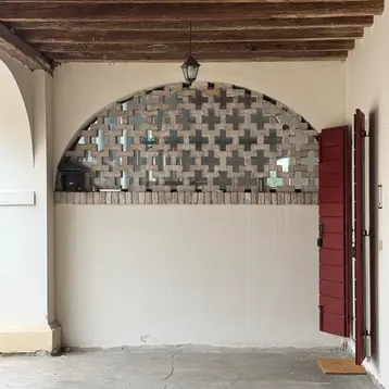 Image of a wall with a round arch. The lower two-thirds are closed and plastered. The upper third of the round arch is filled with glass, and there is also a wall in front of the glass pane. The bricks are deliberately arranged in a gap-filled pattern. The air spaces are arranged in a cross pattern.
