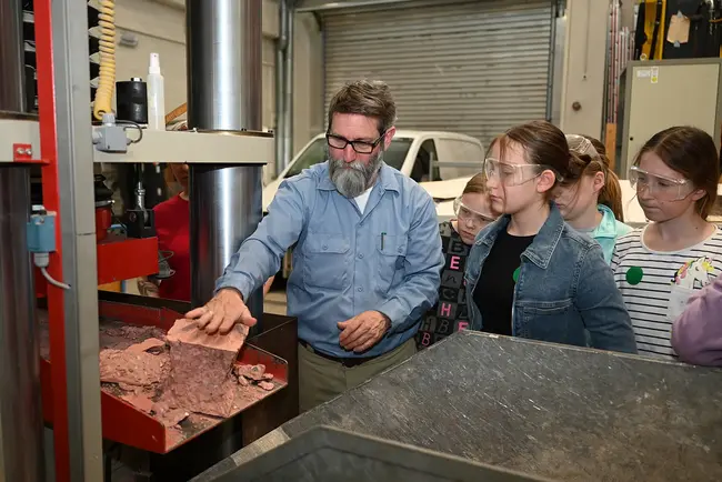 Ein Mitarbeiter der Hochschule RheinMain erklärt Teilnehmerinnen am Girls’Day 2026 eine Maschine im Massivbaulabor. Die Kinder tragen Schutzbrillen.