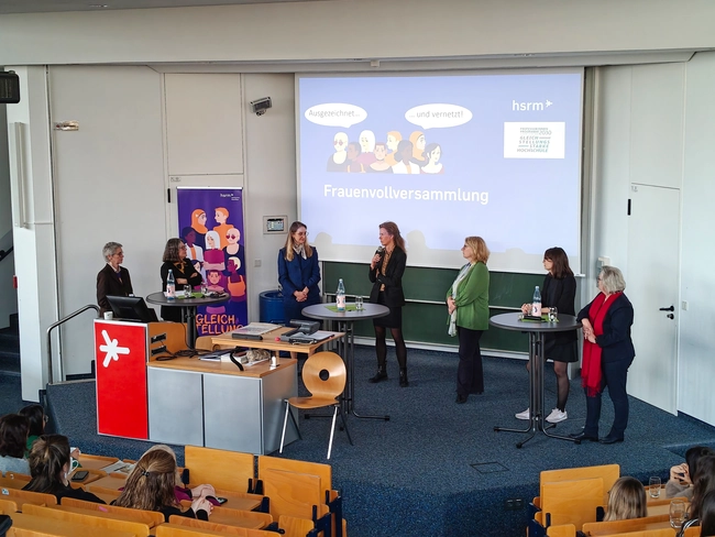 Eine Frauenvollversammlung findet in einem Auditorium statt. Auf der Bühne stehen mehrere Frauen, die miteinander sprechen. Im Hintergrund ist eine Präsentation mit dem Titel „Frauenvolversammlung“ zu sehen, während das Publikum aufmerksam zuhört.