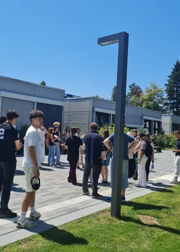 Eine Gruppe von Jugendlichen und jungen Erwachsenen steht draußen in einer Warteschlange vor einem modernen Gebäude. Es ist sonnig, und die Atmosphäre wirkt entspannt. Einige Personen unterhalten sich miteinander.
