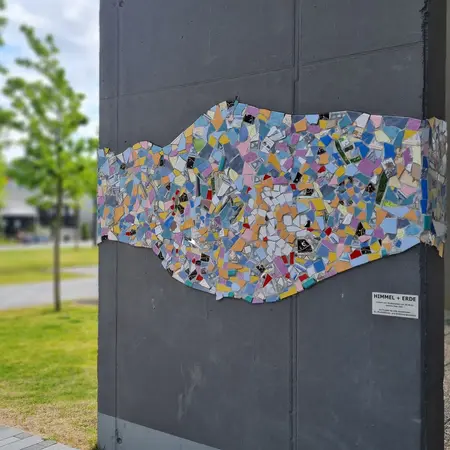 Ein Wandkunstwerk aus bunten Mosaikstücken zeigt eine wellenförmige Struktur. Die Farben variieren von Pastell bis kräftig und bilden ein einzigartiges Muster. Das Kunstwerk befindet sich an einer Gebäudeaußenwand in einem parkähnlichen Umfeld mit Rasen und Bäumen.