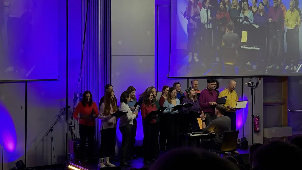 Eine Gruppe von Menschen, die in bunten Oberteilen zusammen singt, steht vor einem Klavier. Der Pianist sitzt mit dem Rücken zur Kamera. Im Hintergrund sind Lichter in lila und blauen Tönen zu sehen. Das Bild vermittelt eine fröhliche und musikalische Atmosphäre.
