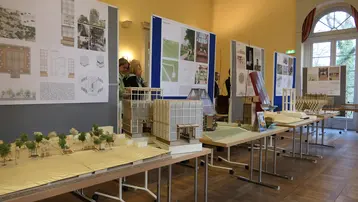 Das Bild zeigt eine Architektur-Ausstellung mit mehreren Tischen auf denen Architekturmodelle stehen, dahinter hängen Poster mit Plänen der Entwürfe.