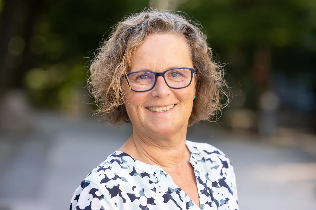 Porträt einer lächelnden Frau mit lockigem, braunem Haar und einer blauen Lesebrille. Sie trägt eine blumige Bluse und steht im Freien mit einem verschwommenen Hintergrund aus Bäumen und Gehwegen.